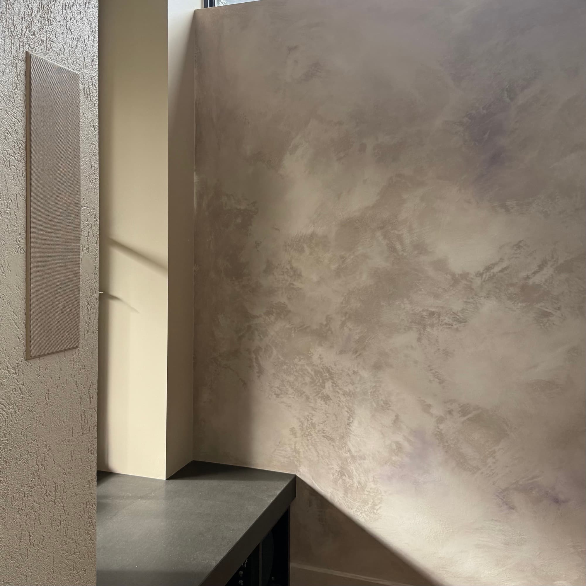 A hand-finished mineral wall with low sunlight crossing it, beside a textured render return and a dark stone bench.